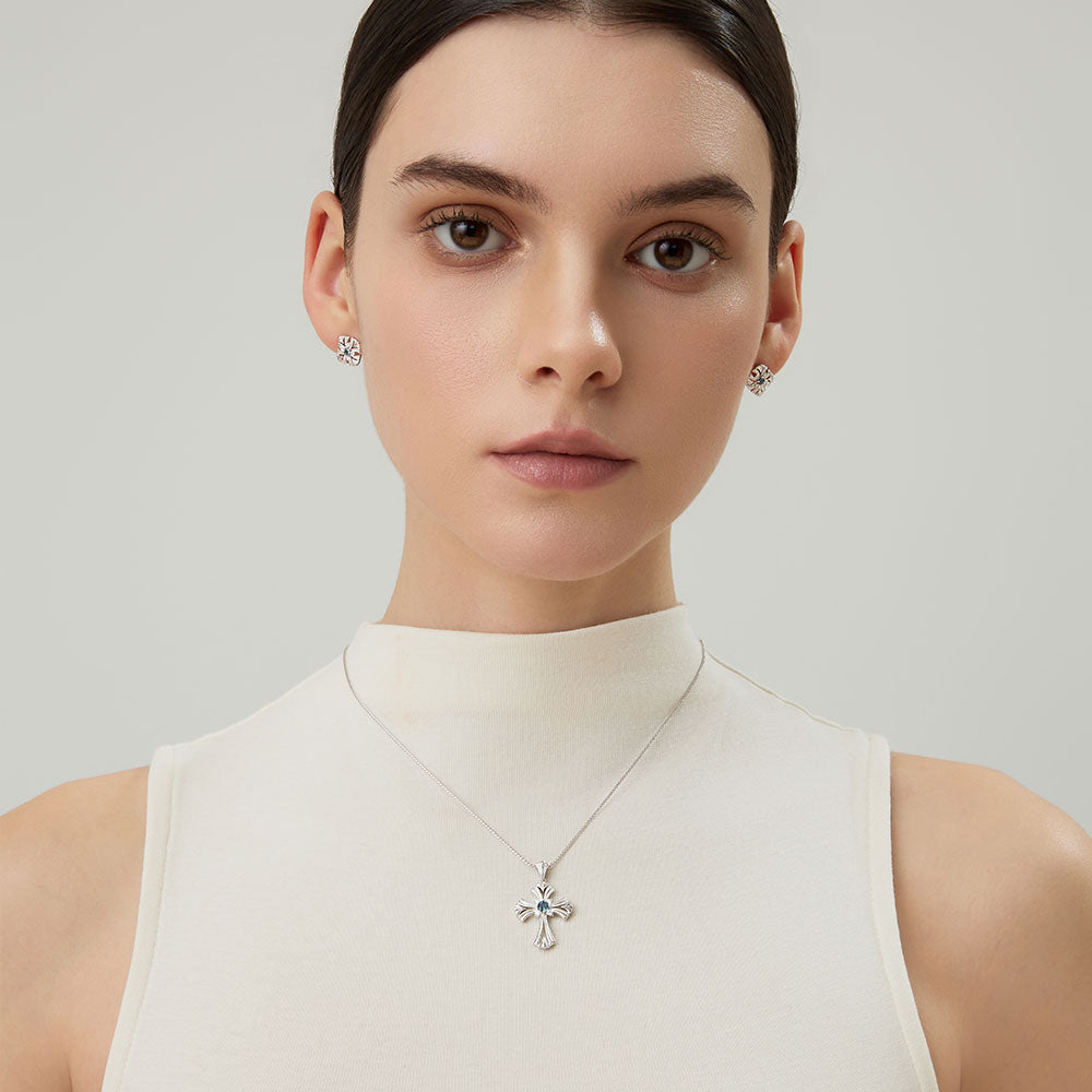 Fashion model wearing TIKOO original design blue topaz cross necklace with a white mock neck top.