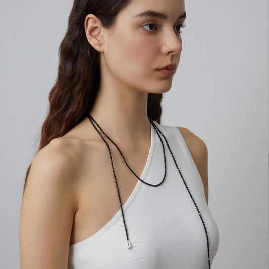 Professional model showcasing the versatile TIKOO black spinel beaded lariat necklace, styled with a minimalist white one-shoulder top.