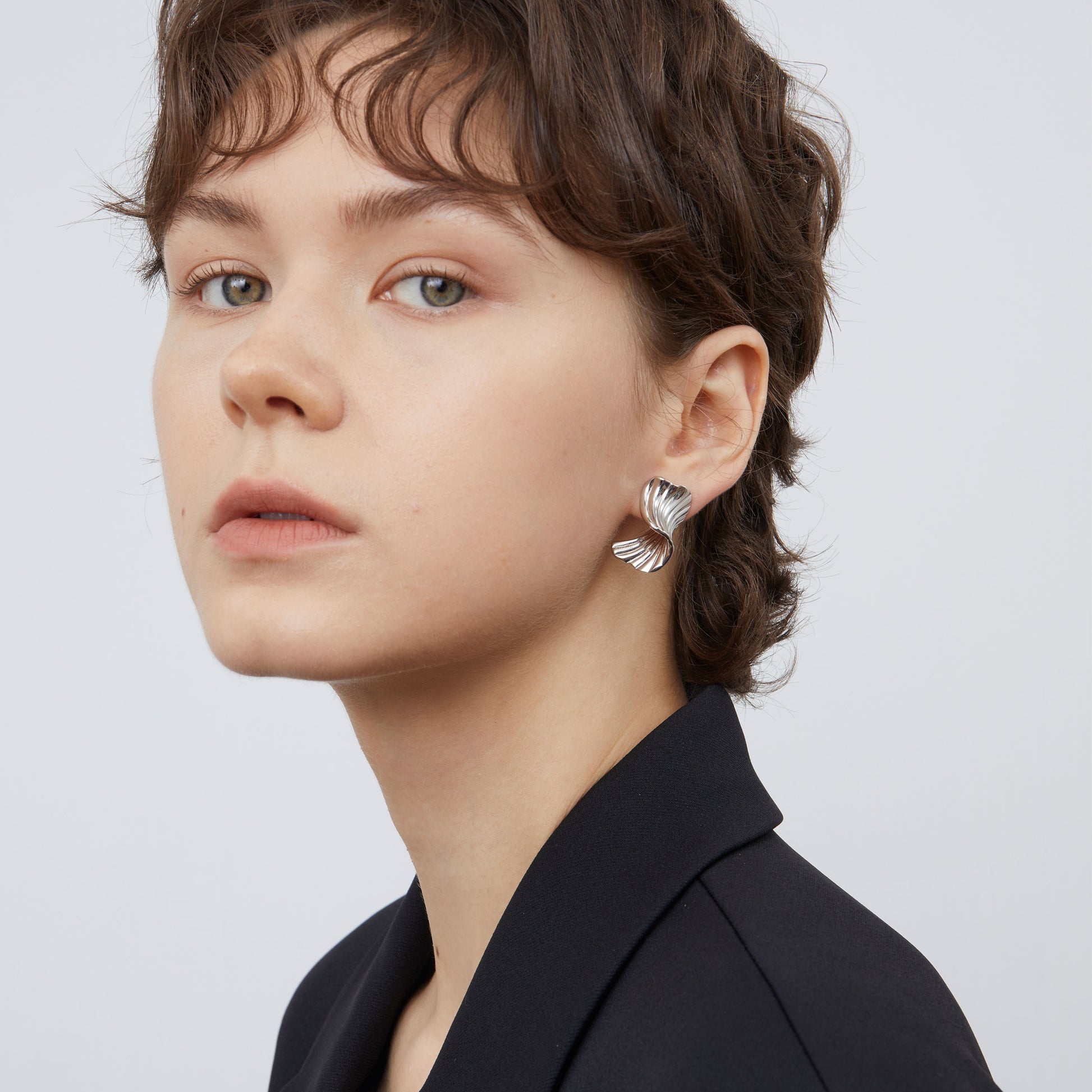 Side view of a model wearing TIKOO white gold plated sculptural earrings with a modern black blazer.